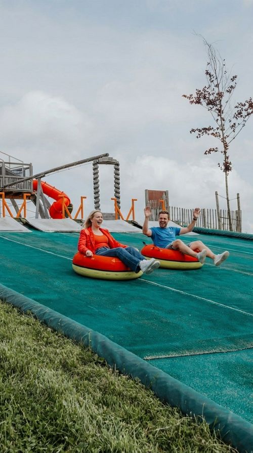 IKUNA Triple Tubing Bahn Zwei lächelnde Erwachsene in Rot und Blau sausen auf aufblasbaren Reifen die IKUNA Triple Tubing Bahn unter bewölktem Himmel hinunter.