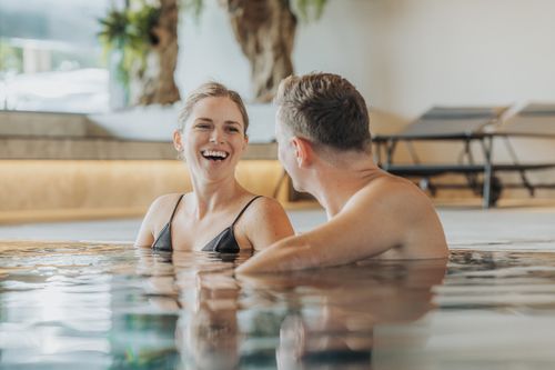Eine lachende Frau und ein Mann stehen einander im Indoorpool der IKUNA Spa- & Wellnesswelt gegenüber. Im Hintergrund befinden sich Liegen und Pflanzen.