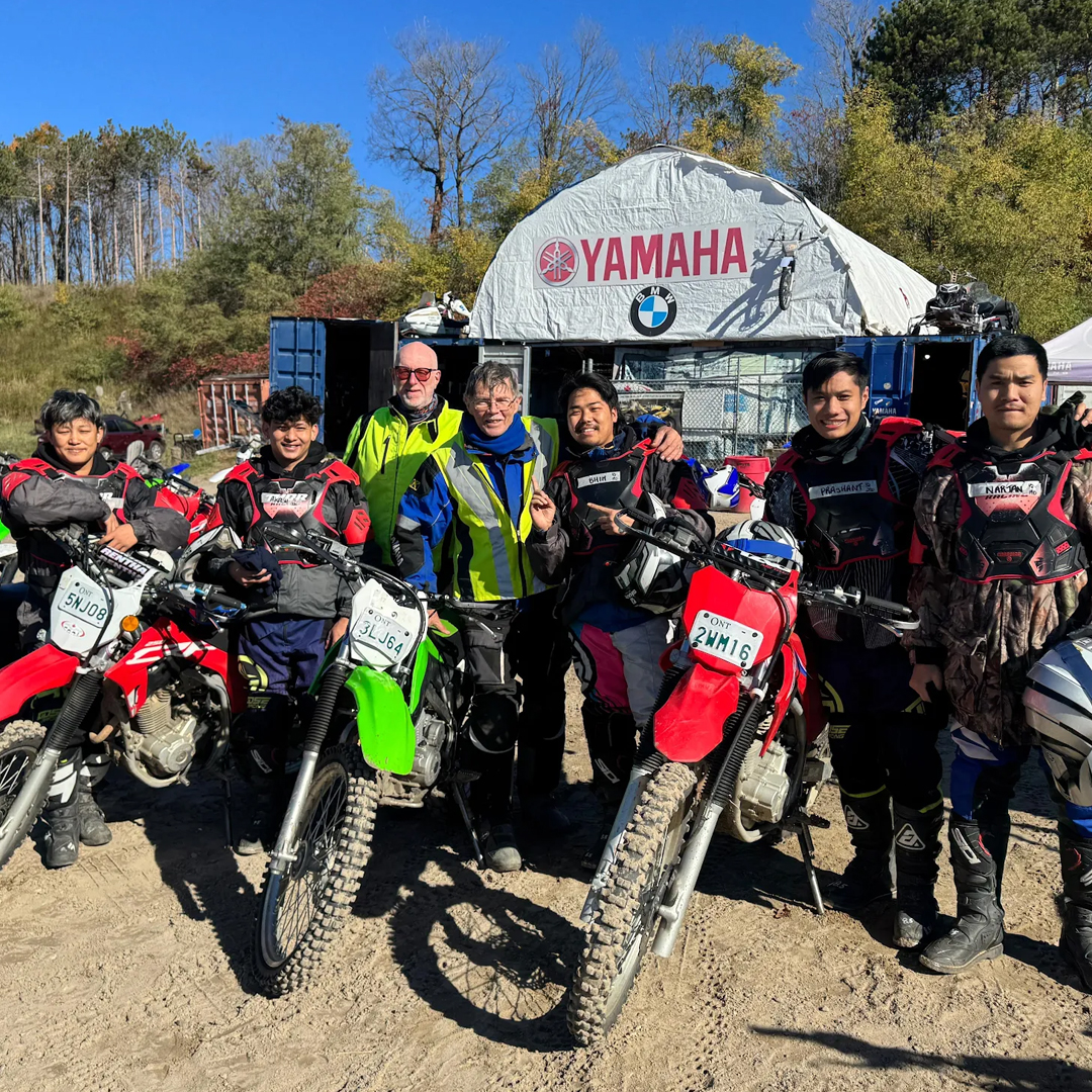 Group of people in motorcycle gear pose with dirt bikes in front of a Yamaha tent at an outdoor event.