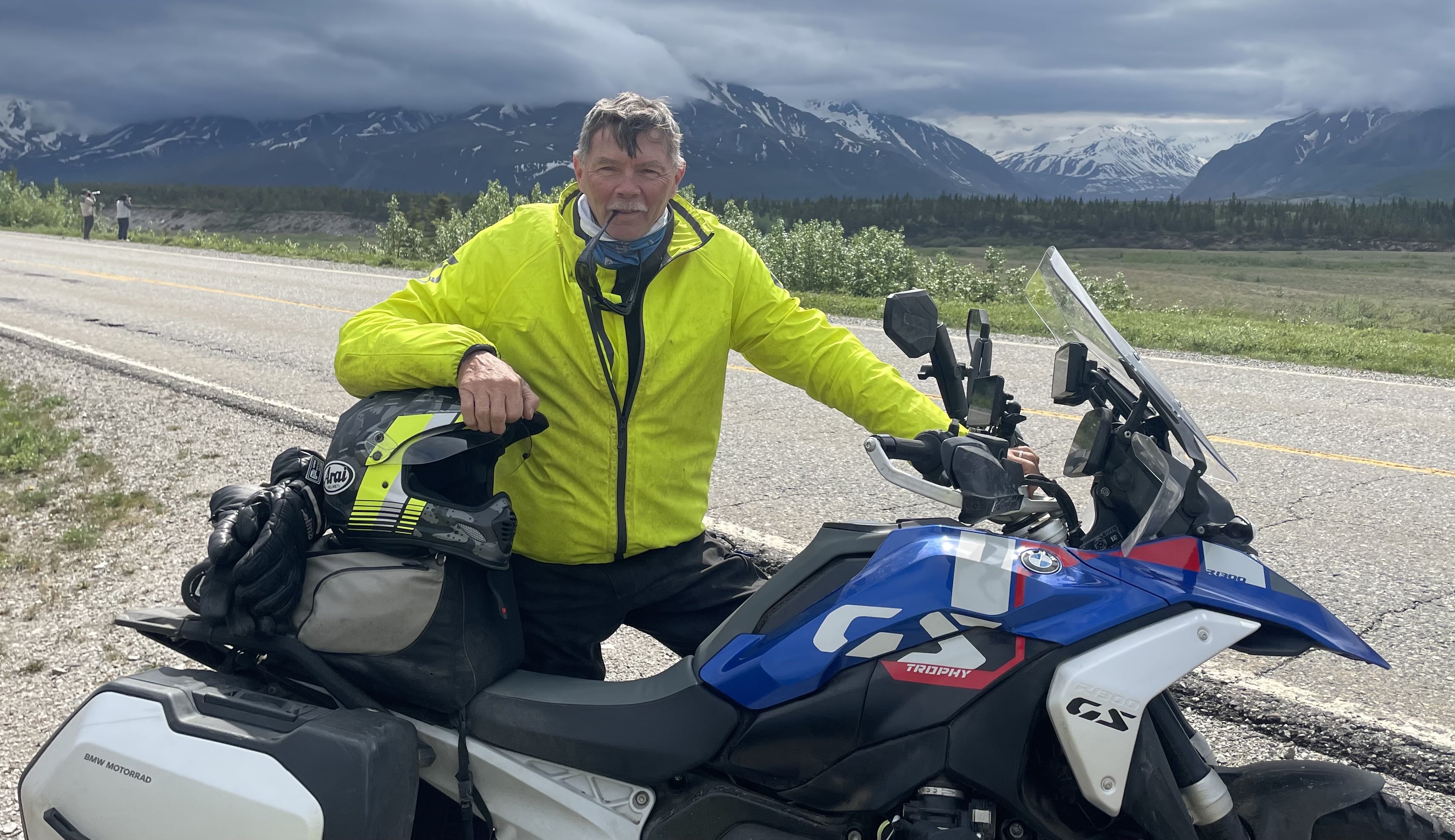 Man in a bright yellow jacket stands beside a blue and white motorcycle on a scenic road with mountains and cloudy skies in the background.