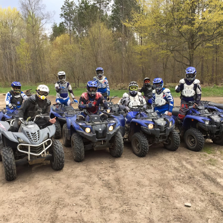 A group of people in helmets and gear sit on ATVs in a forested area, ready for an off-road adventure.