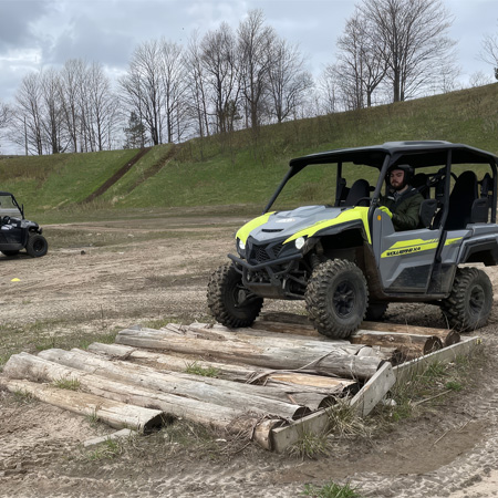 An off-road vehicle drives over a log bridge on a muddy terrain with bare trees in the background. Another vehicle is visible in the distance.