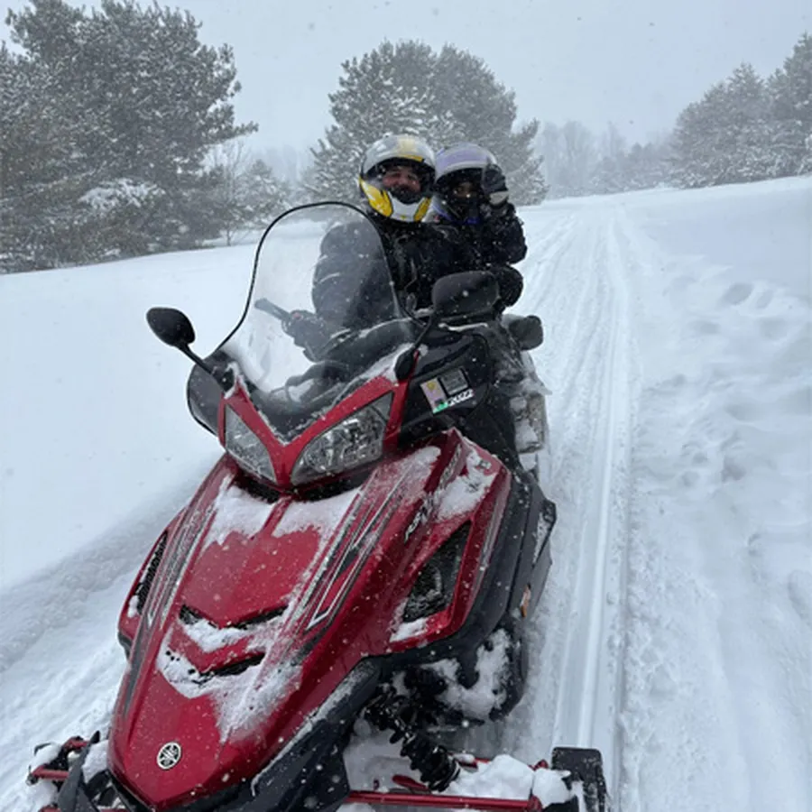 Two people riding a red snowmobile on a snowy forest trail.