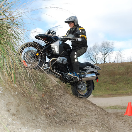 Rider in protective gear climbs a steep dirt hill on a motorcycle, with orange traffic cones marking the path.
