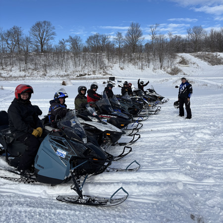 A group of people in winter gear sitting on snowmobiles lined up on a snowy landscape, with one person standing nearby. Trees in the background.