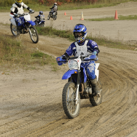 Motorcyclists in blue and white gear riding dirt bikes on a sandy track, with traffic cones marking areas in the background.