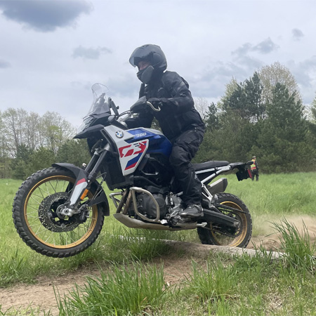 Motorcyclist in black gear riding a blue and white bike on a grassy trail, with trees and a cloudy sky in the background.