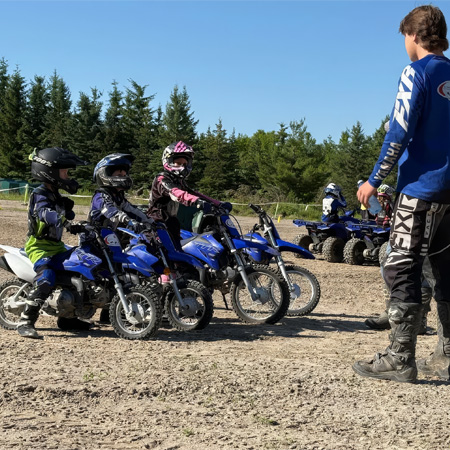 Children on blue dirt bikes line up on a dirt track, guided by an adult instructor. Trees are visible in the background.