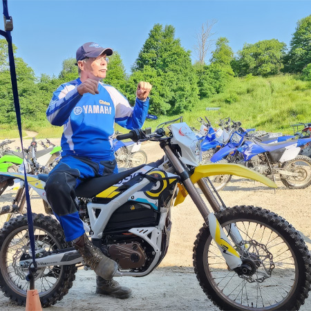 A person in a Yamaha jersey sits on a dirt bike, gesturing energetically. Several other bikes are parked nearby on a sunny day with greenery.