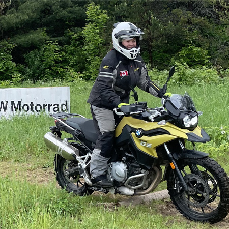 Person wearing motorcycle gear and helmet rides a yellow BMW GS motorcycle on a grassy trail near a BMW Motorrad sign.