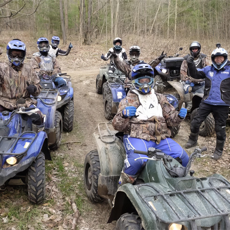 A group of people wearing helmets and protective gear are posing with their ATVs on a forest trail.