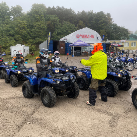 Group of people in helmets and gear sitting on blue ATVs with an instructor in a bright jacket at an outdoor venue with a Yamaha tent.