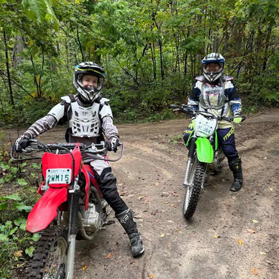 Two dirt bike riders stopped on a wooded trail, facing the camera.
