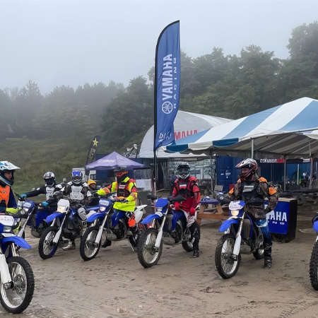 A group of motorcyclists in gear sit on Yamaha dirt bikes, lined up in front of tents and flags on a foggy day.