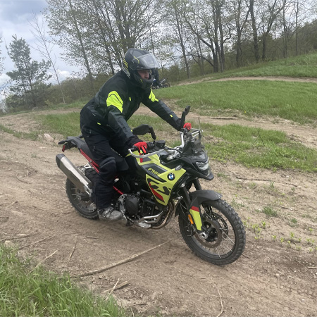 Rider in black and neon yellow gear on a BMW motorcycle navigating a dirt trail surrounded by grass and trees.