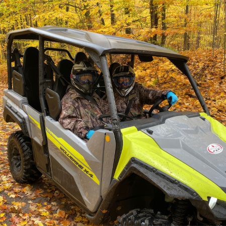 Two people in camouflage gear and helmets sitting in a green and gray ATV, surrounded by vibrant autumn foliage on a trail.