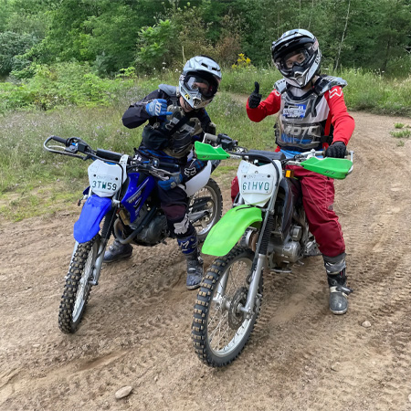 Two dirt bikers on off-road bikes give thumbs up, wearing helmets and gear, surrounded by greenery.
