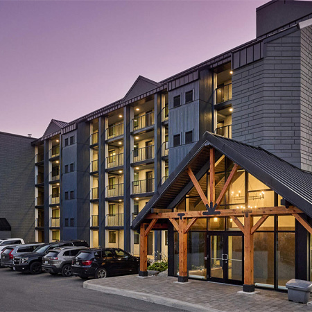 Modern multi-story hotel with a gabled entrance, illuminated windows, and parked cars at dusk under a purple sky.