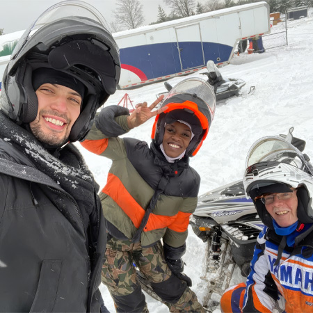 Three people wearing helmets and winter gear stand smiling on a snowy field next to a snowmobile.