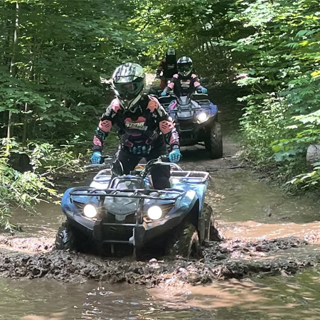 ATV riders navigate a muddy forest trail, wearing protective gear and helmets, surrounded by lush green foliage.