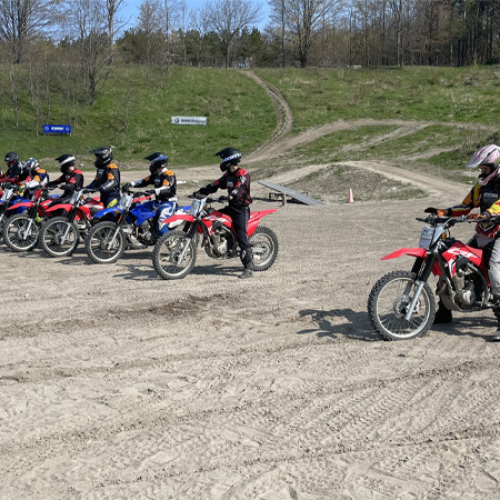 Motorcyclists in colorful gear line up on dirt bikes at an outdoor motocross track, surrounded by trees and grassy areas.