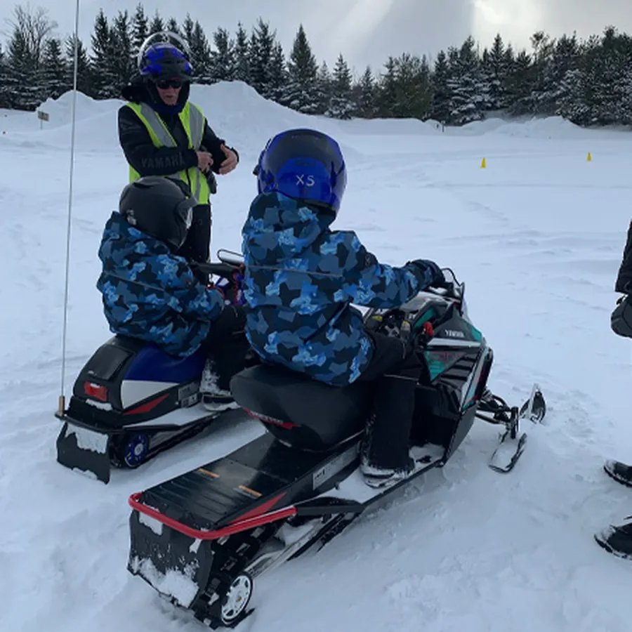 Two children riding a snowmobile with an instructor in a snowy area.
