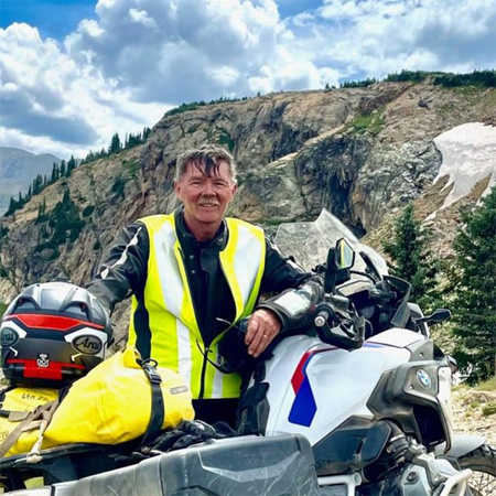Man in a high-visibility jacket stands by a loaded motorcycle in a mountainous area, with rocky cliffs and clouds in the background.
