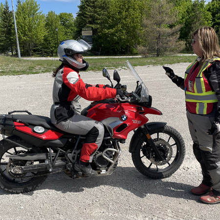 Two people in motorcycle gear; one is seated on a red motorcycle, and the other is standing and gesturing, in an outdoor setting.