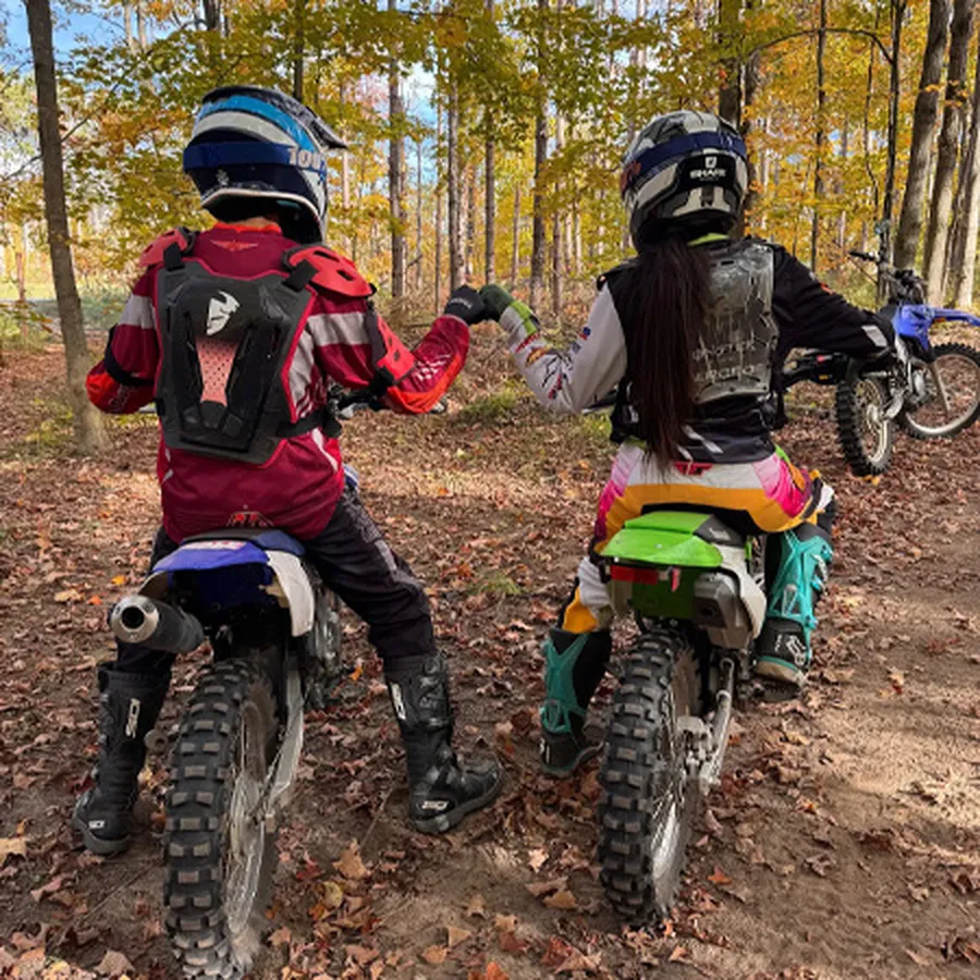 Two dirt bike riders stopped on a forest trail, fist bumping each other.