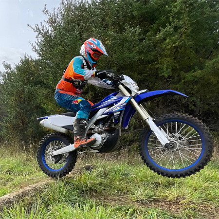 A person on a dirt bike wearing colorful gear, riding over grass with trees in the background.