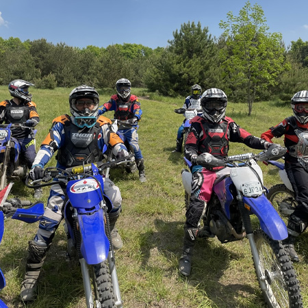 A group of dirt bikers wearing helmets and riding gear on blue motorcycles, gathered on a grassy field under a clear sky.