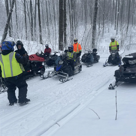 A group of people on snowmobiles gather on a snowy trail in a forest, wearing colorful jackets and helmets.