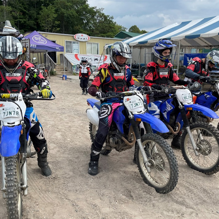 Group of motocross riders in colorful gear and helmets, lined up on dirt bikes in front of tents and a small building.