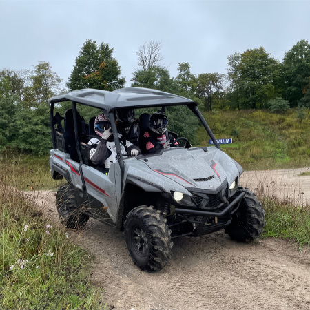 Two people wearing helmets ride in a rugged, off-road vehicle on a dirt path, surrounded by greenery and overcast skies.