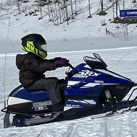 Child in a helmet rides a blue and white snowmobile on snowy terrain, surrounded by trees and a sign in the background.