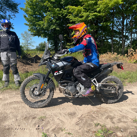 Person in colorful riding gear on a BMW GS off-road motorcycle, with another person standing nearby on a dirt path surrounded by trees.