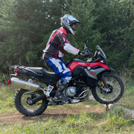 Motorcyclist in full gear riding a red BMW GS off-road on a dirt path, surrounded by green trees.