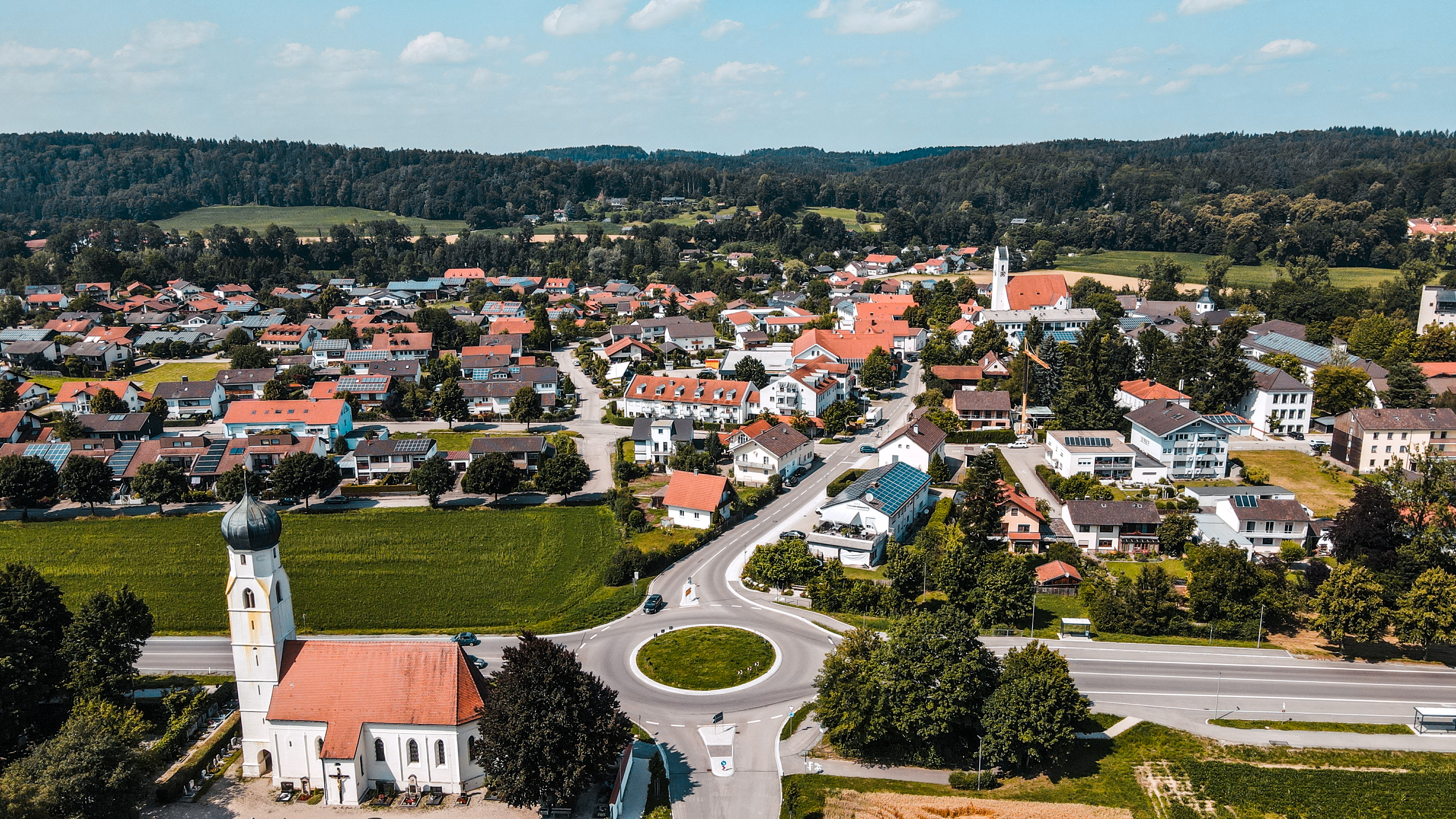 Luftaufnahme der Gemeinde mit Kirche, Kreisverkehr und Wohngebiet. Rote Ziegeldächer, Grünflächen und landwirtschaftlich genutzte Felder prägen das Ortsbild vor einer bewaldeten Hügellandschaft.