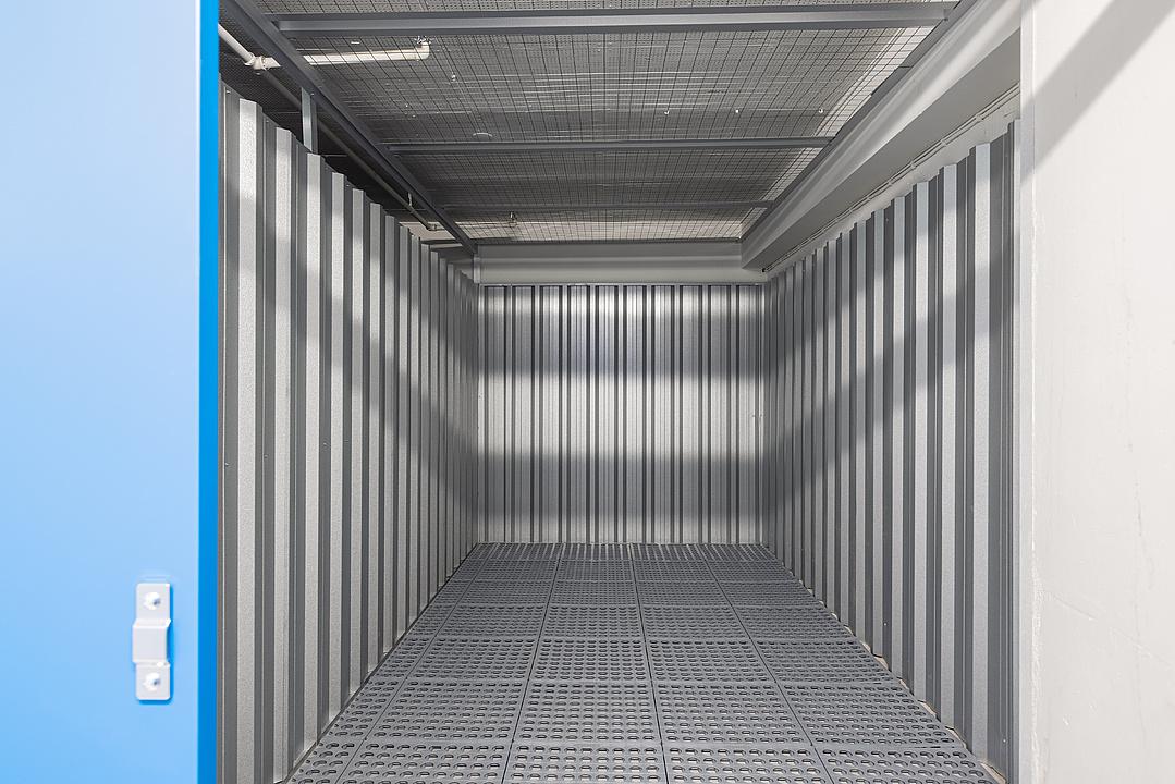 Empty metal storage unit with open blue door, featuring corrugated walls, grid floor, and a mesh ceiling.