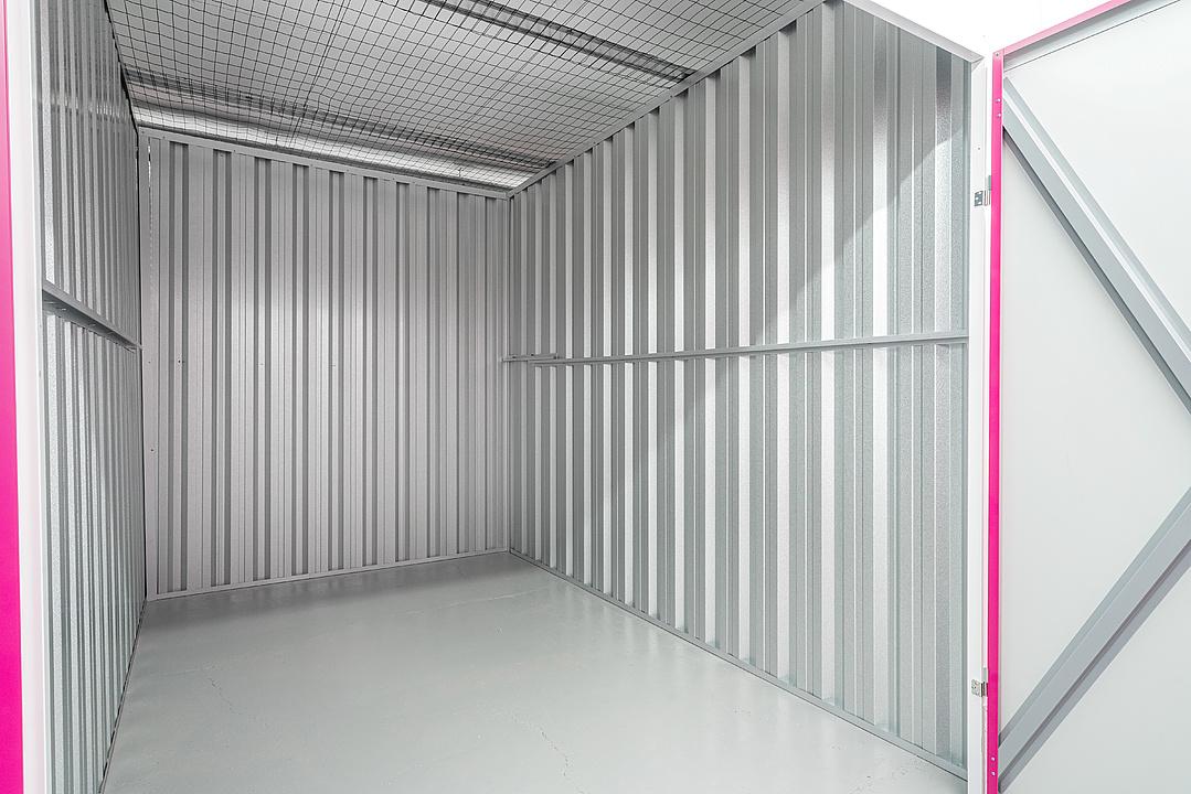Empty metallic storage unit with corrugated steel walls and a wire mesh ceiling. The open door features a bright pink frame.