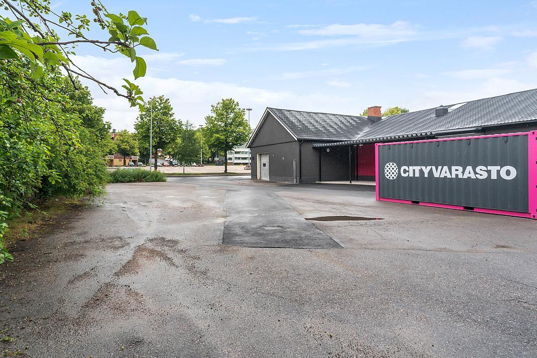 Paved lot with wet patches, surrounded by greenery. A gray building and a pink-trimmed storage container labeled "CITYVARASTO."