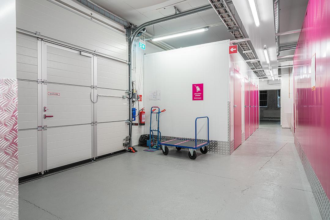 Indoor storage space with pink lockers, a large closed door, a blue handcart, and overhead lighting on a concrete floor.