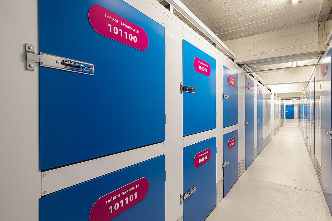 A corridor of blue storage unit doors, each labeled with a pink plaque containing a unique number and text. The floor and ceiling are white.