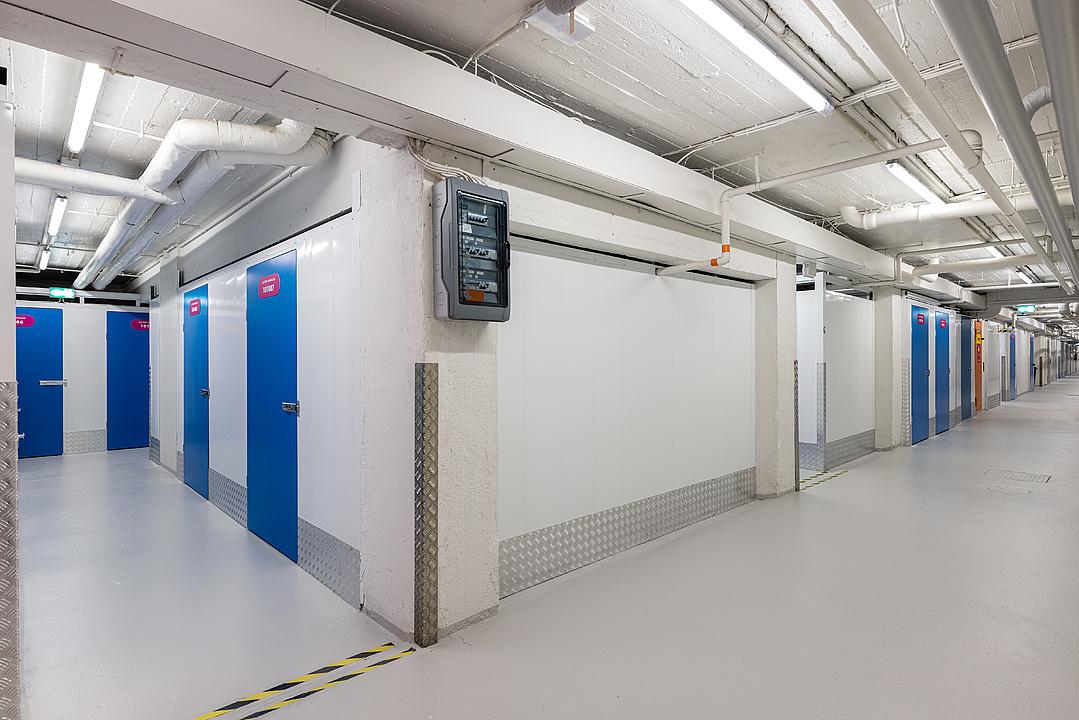 A clean, well-lit storage facility hallway with blue doors, white walls, visible pipes, and an electrical panel on the wall.
