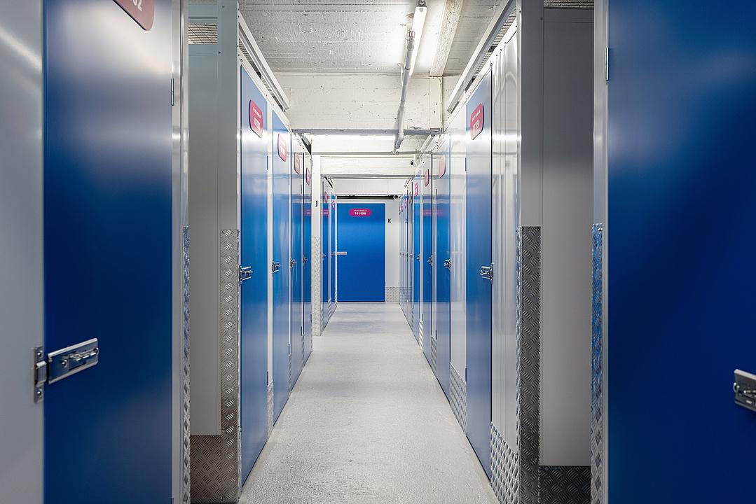 Hallway with rows of blue metal doors on both sides, leading to a closed door at the end. Bright fluorescent lighting illuminates the space.
