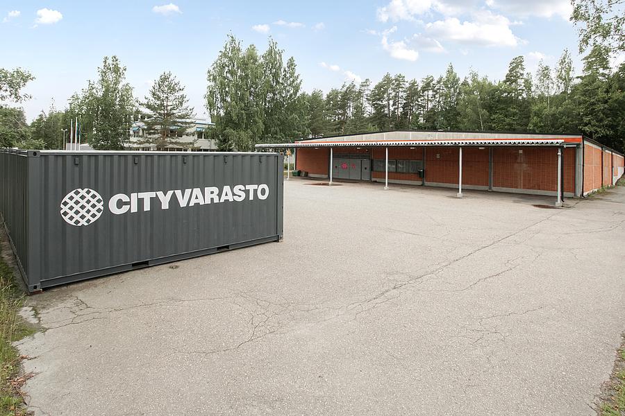 A storage facility with a large container labeled "Cityvarasto" in an open paved area, surrounded by trees and a brick building.