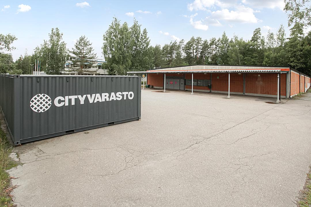 A storage facility with a large container labeled "Cityvarasto" in an open paved area, surrounded by trees and a brick building.