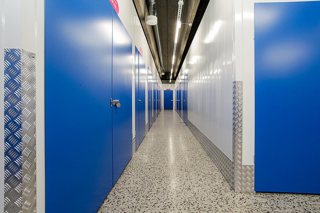 A well-lit corridor in a storage facility, featuring rows of blue metal doors with locks and a polished speckled floor.