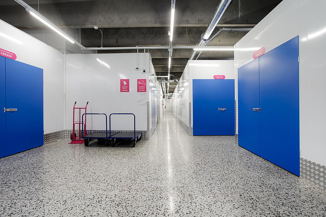 Interior of a storage facility with polished concrete floor, blue doors, and two hand trucks. Bright overhead lighting illuminates the corridor.