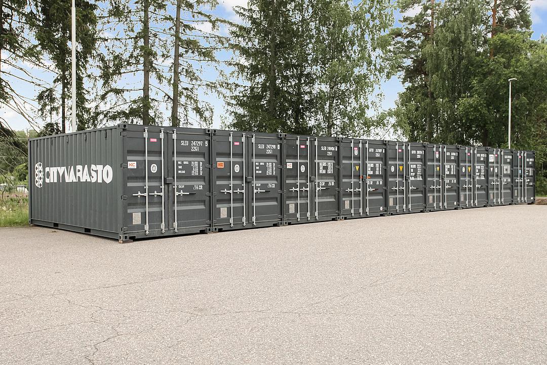 A row of dark gray shipping containers with "CITYVARASTO" branding, parked on an asphalt lot, surrounded by trees.
